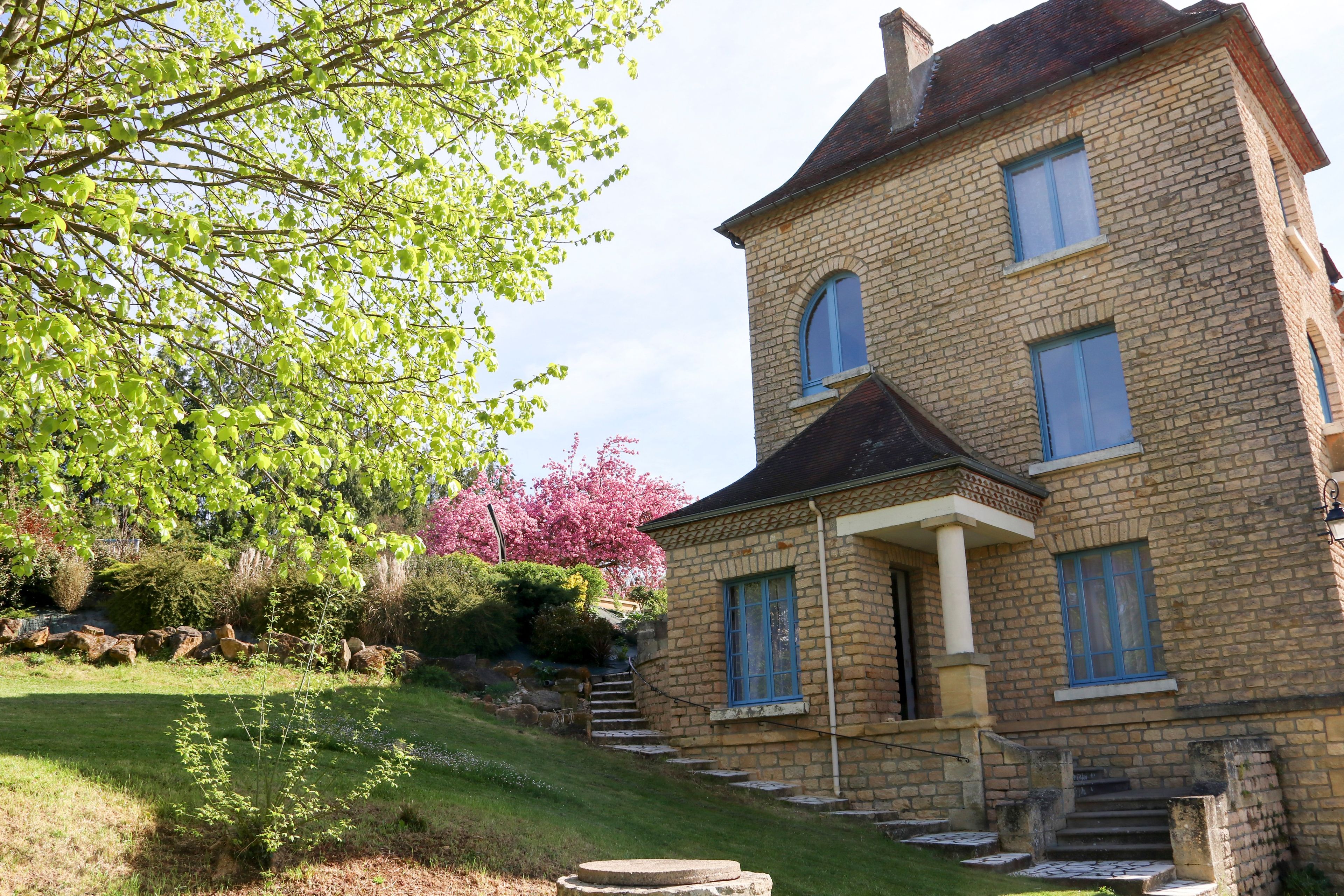 Un bâtiment en pierre de deux étages, de style traditionnel, avec une entrée principale accessible par des marches, un petit porche et une fenêtre bien visible, dotée de volets bleus.