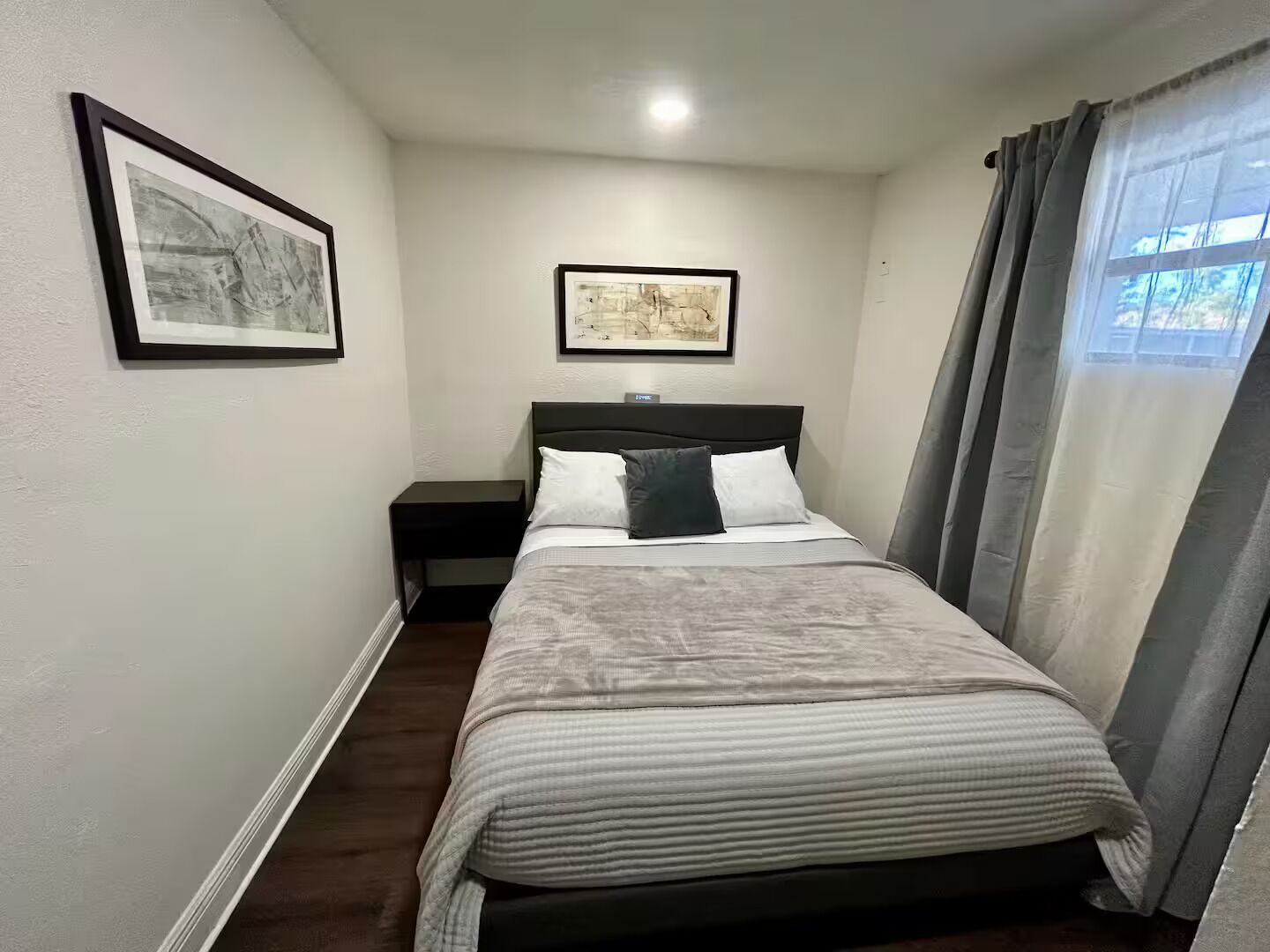 Un dormitorio con cama, una mesita de noche, una lámpara de pared y dos cuadros enmarcados.