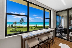 A room with a large window overlooking a beach and ocean, a bar area with stools, and a dining table with chairs. - Wake up to breathtaking direct Gulf-front views at Loggerhead Cay 142. This beautifully updated 2-bedroom, 2-bath beachfront condo sleeps 5 and includes beach gear, a heated pool, pickleball, and everything you need for the perfect Sanibel getaway. (Sanibel Island)