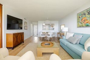Un salon moderne avec un canapé, une table basse et un téléviseur. On aperçoit à l’arrière-plan un coin repas avec des chaises et une table.