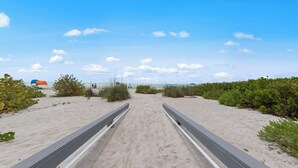 En strandpromenad som leder ner till havet, omgiven av vegetation och en klar himmel.