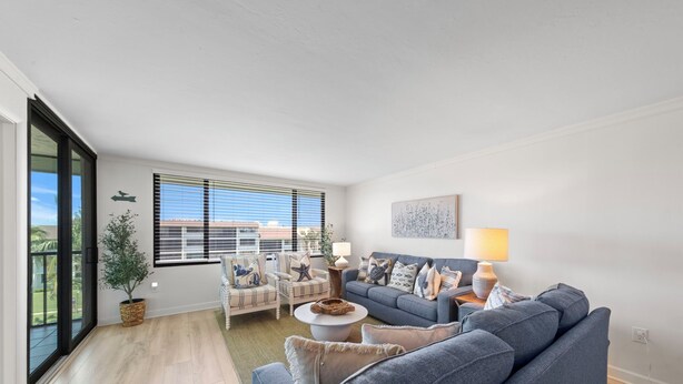 Un salon moderne avec un canapé gris, un fauteuil blanc et une table basse. On y trouve une grande fenêtre avec des stores, une plante en pot et une lampe fixée au mur.