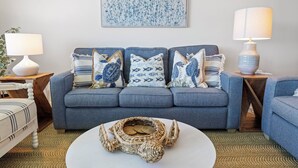 Un salon avec un canapé gris, des coussins à motifs, un panier en osier posé sur une table basse blanche et un tableau accroché au mur.