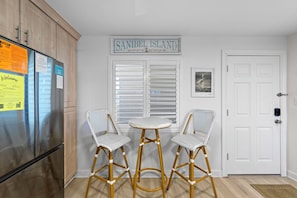 A kitchen with wooden cabinets, a refrigerator, a sign reading 'Sanibel Island,' and two wicker chairs with a small table. - Wake up to Gulf views at Loggerhead Cay 584, a top-floor 2BR/2BA Sanibel condo overlooking the pool with beautiful side Gulf views + additional water views from the guest bedroom. Enjoy direct beach access, heated pool, tennis/pickleball and more (Sanibel Island)