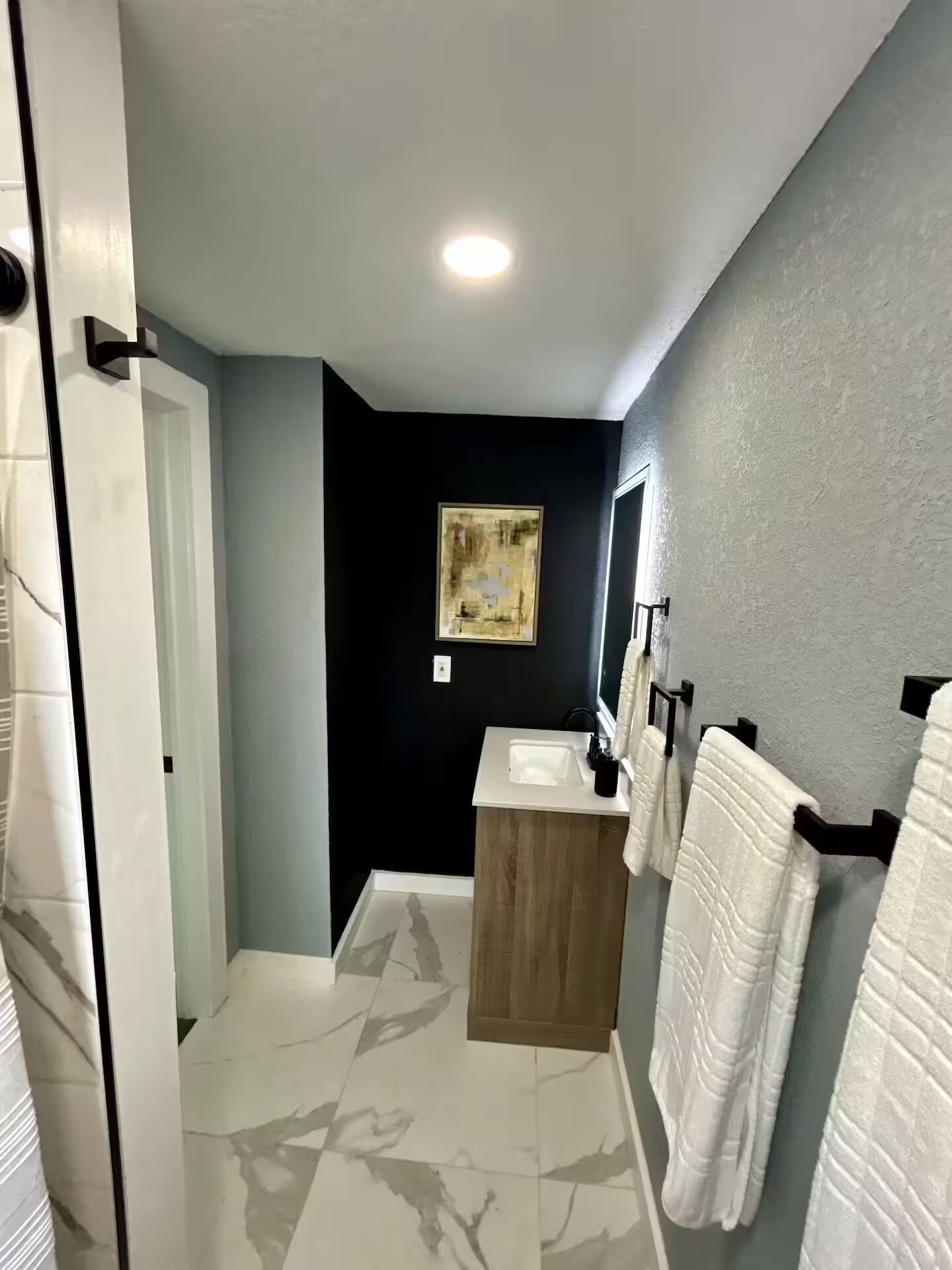 A bathroom with a white sink, a wooden vanity, a black accent wall, and white towels hanging on hooks.