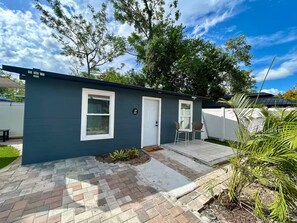 A single-story house with a dark exterior, a white door, and a small porch with two chairs. - Sunshine Guest Cottage (Tampa)