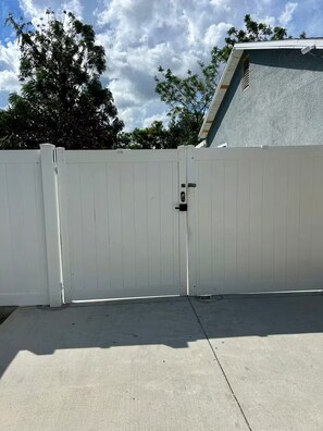 A white metal gate with a lock, leading to a building with a grey roof and a white fence. - Sunshine Guest Cottage (Tampa)