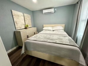 A bedroom with a bed, a dresser, an air conditioner, and a window with curtains. - Sunshine Guest Cottage (Tampa)