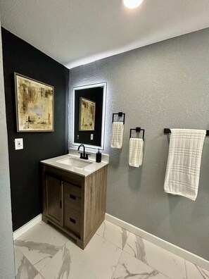 A bathroom with a dark wall, a white sink, a mirror, and two towels. - Sunshine Guest Cottage (Tampa)