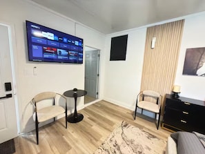 Ein modernes Hotelzimmer mit einem wandmontierten Flachbildfernseher, der verschiedene Streaming-Dienste anzeigt. Zwei Sessel und ein kleiner runder Tisch mit einer Pflanze stehen im Raum. Der Boden ist mit hellem Holz versehen und es gibt ein großes Fenster mit Jalousien.