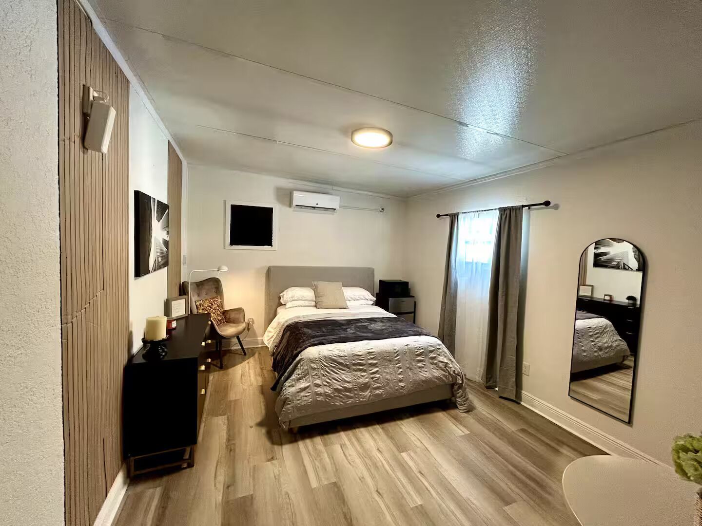 A bedroom with a bed, a desk, a chair, a mirror, and an air conditioning unit.