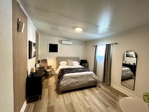 Ein Schlafzimmer mit einem Bett, einem Schreibtisch, einem Sessel, einem Spiegel und einer Klimaanlage.