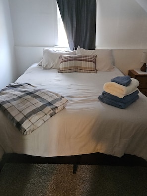 A bed with a white bedspread, two pillows, a plaid blanket, and a stack of folded towels.