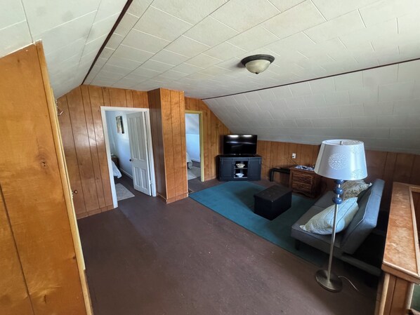 Ein Zimmer mit schräg laufender Decke, Holzwänden, einer Couch, einer Lampe, einem Fernseher und einem kleinen Schrank.