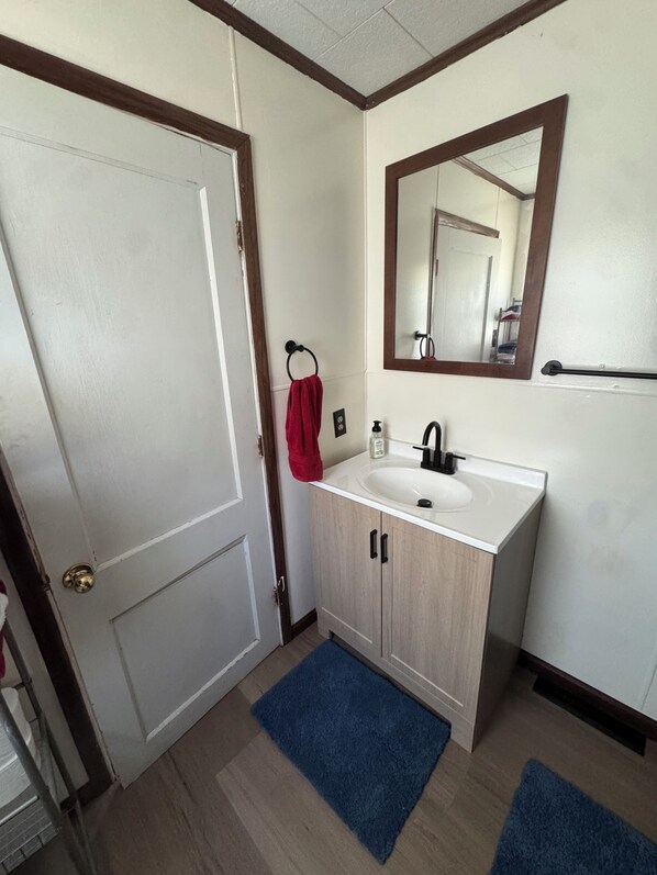 Un baño con una puerta blanca, un tocador de madera con un lavabo, un espejo y una alfombra azul en el suelo.