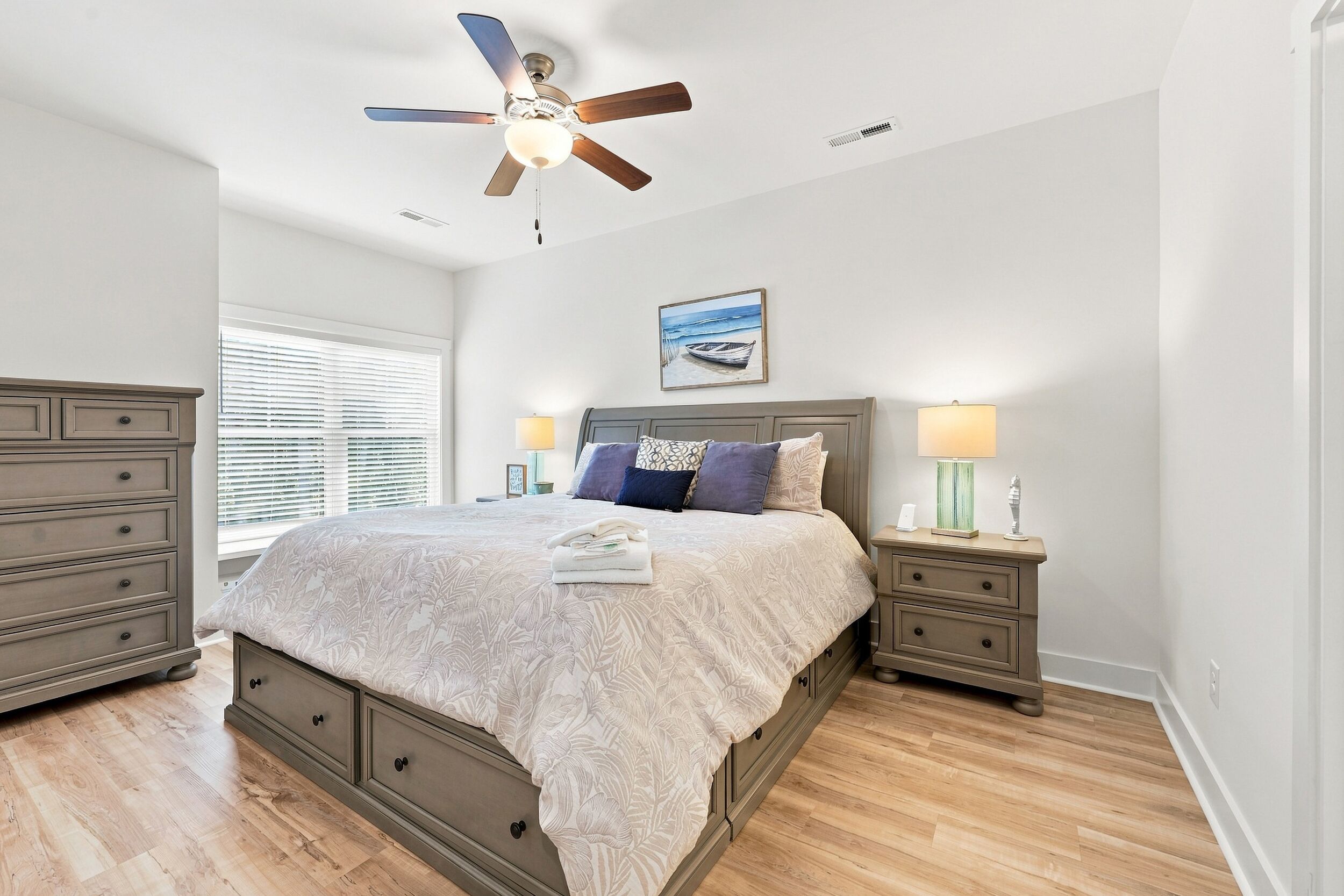 A bedroom with a bed, nightstands, a ceiling fan, a window with blinds, and a painting on the wall.