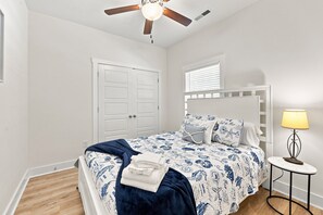 Ein Schlafzimmer mit Bett, Nachttisch, Lampe, Deckenventilator und einem Fenster mit Jalousien.