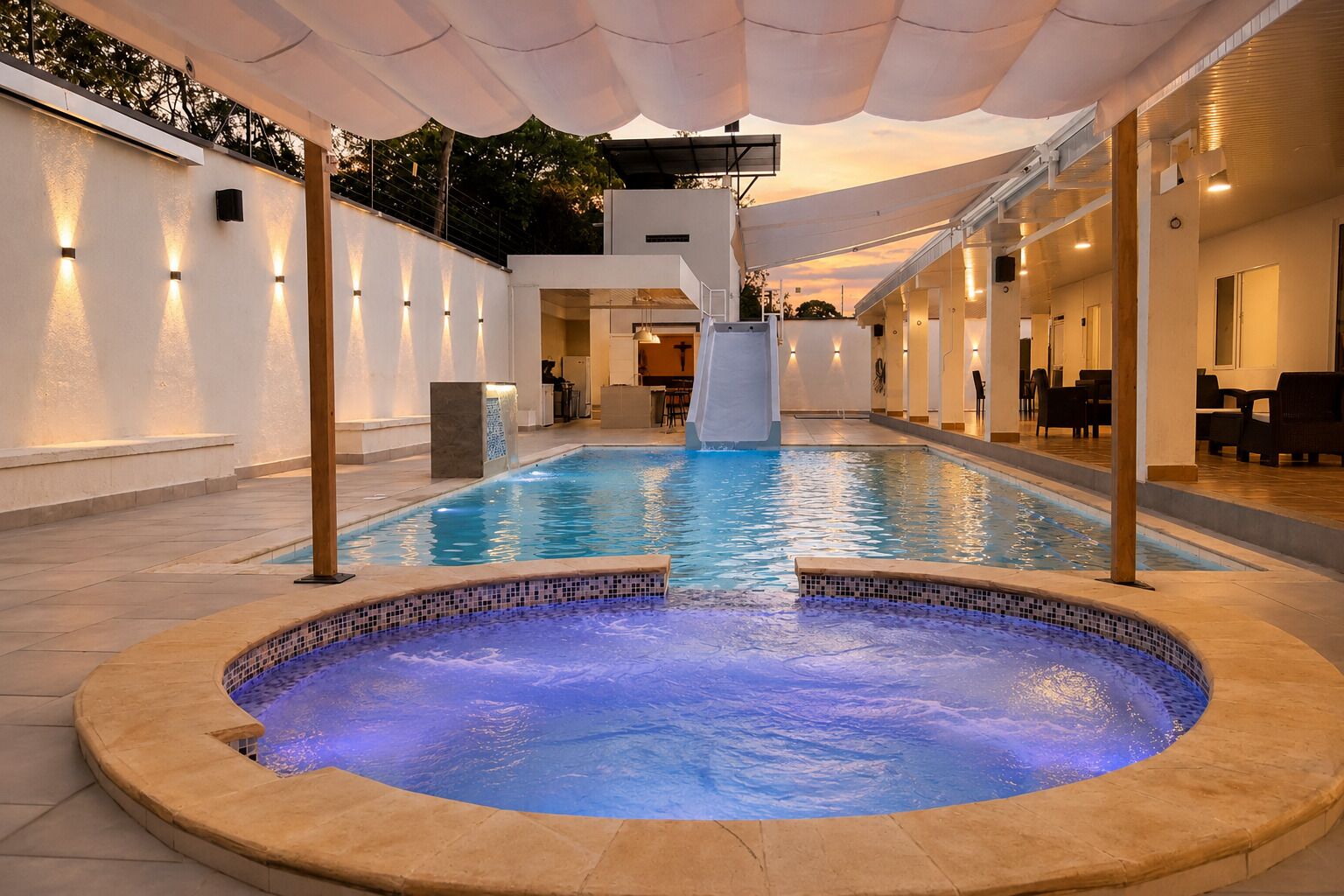 Une piscine intérieure avec un jacuzzi, entourée d’un espace couvert aménagé avec des sièges et offrant une vue sur l’extérieur.