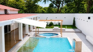 A swimming pool with a wooden deck, a covered seating area, and a white building with a red roof.