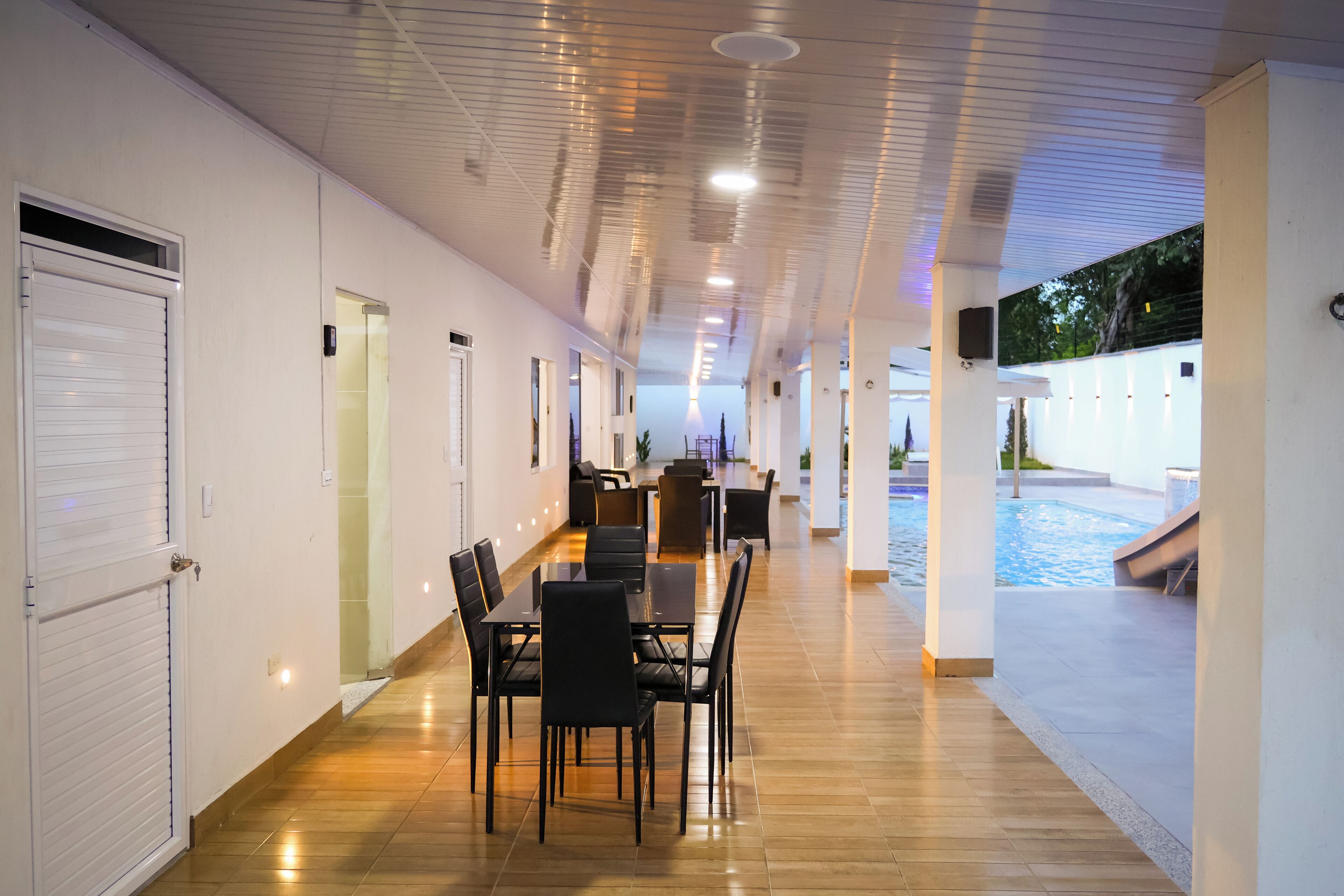Un couloir donnant sur un espace piscine, un coin repas avec des tables et des chaises, et un mur extérieur blanc.