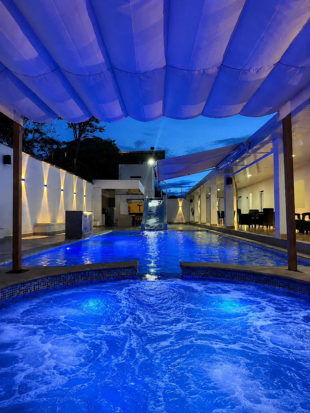 Une piscine avec un toboggan, entourée d’espaces de repos couverts et d’un bâtiment à l’architecture moderne.