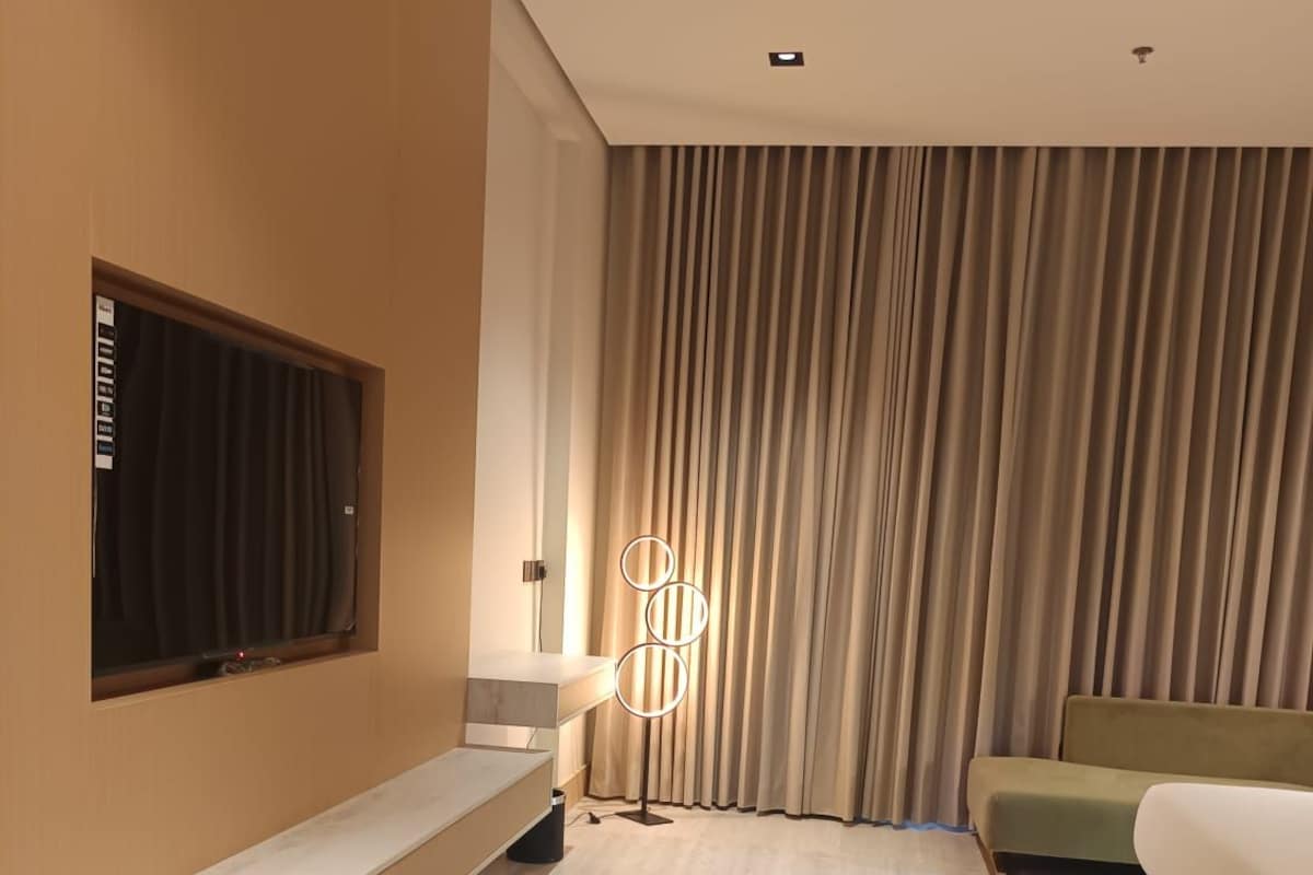 A modern hotel room with a large window, a flat-screen TV, a sofa, a bedside table, and a floor lamp.