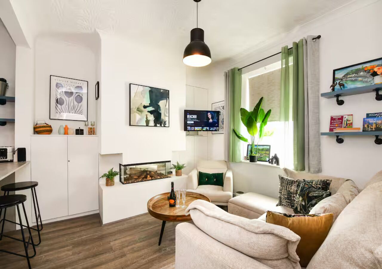 A modern living room with a sofa, coffee table, and a TV. There is a kitchen area with a bar stool and a plant.