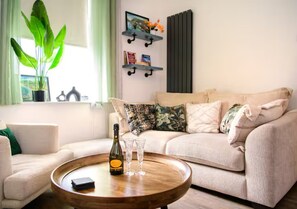 A living room with a beige sofa, a wooden coffee table, a bottle of wine, and two glasses. There is a plant on the left and a wall-mounted shelf with books and decorative items. - Modern 2BR Cardiff Home | 2 Bath, Close to Centre - Pass the Keys (Cardiff)