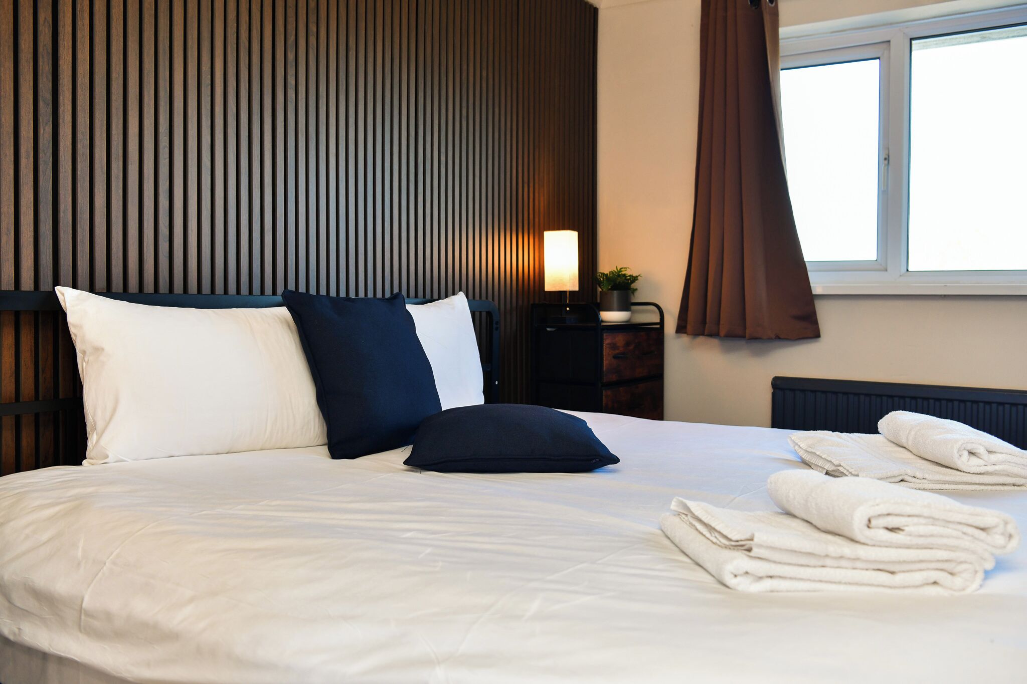 A neatly made bed with pillows, a bedside lamp, a window with curtains, and folded towels on the bed.