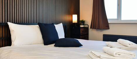 A neatly made bed with pillows, a bedside lamp, a window with curtains, and folded towels on the bed.