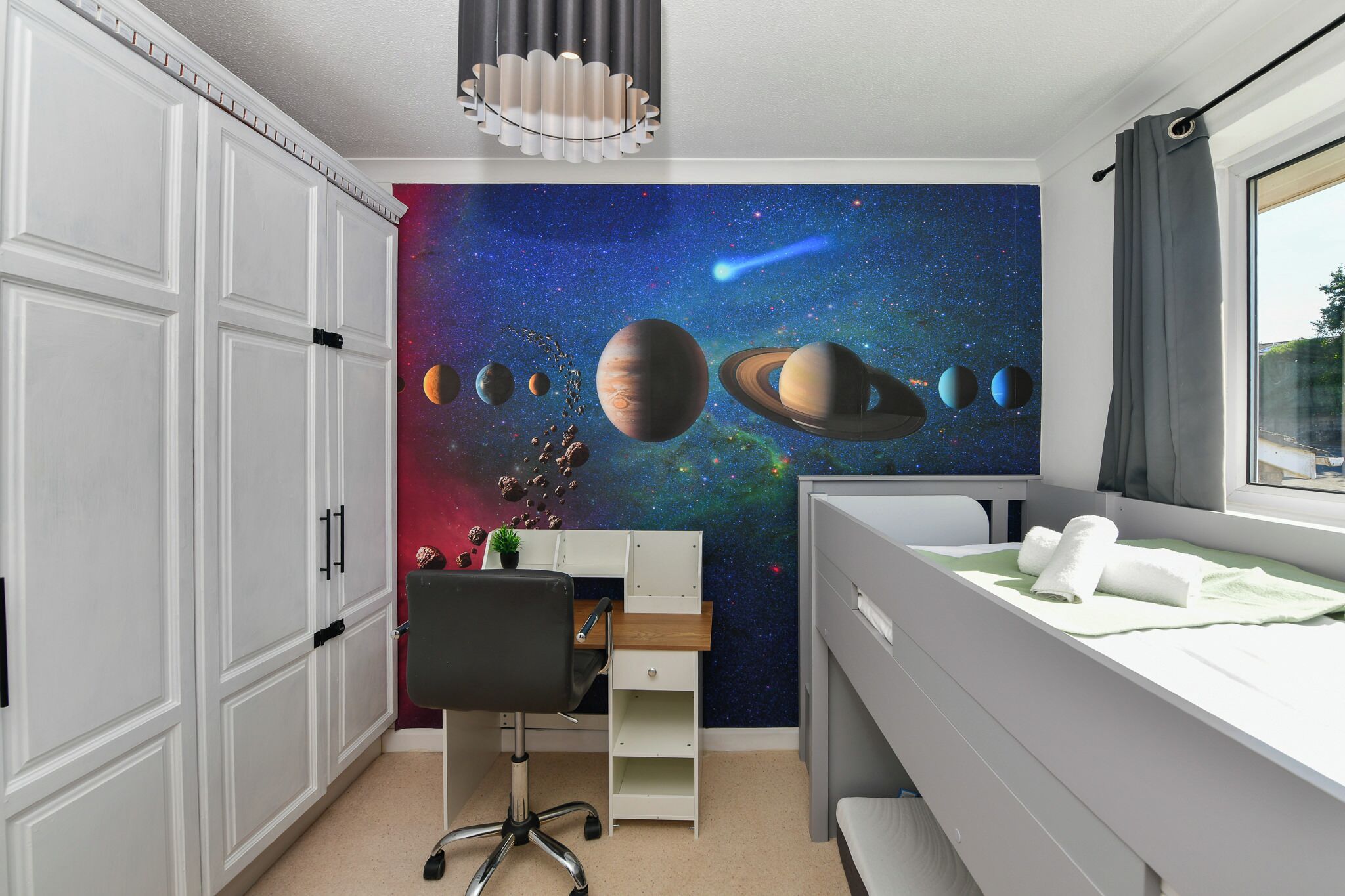 A bedroom with a space-themed wall mural, a desk with a chair, a bed, and a window with curtains.