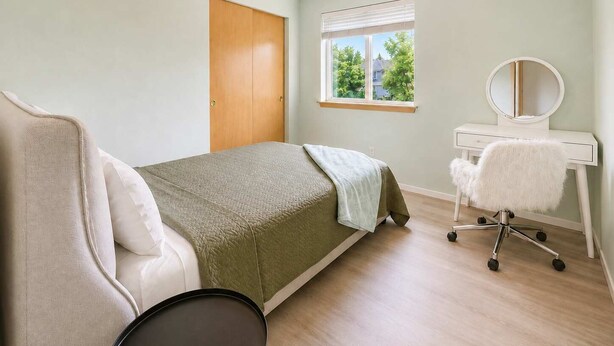 Quarto com cama, escrivaninha com espelho, cadeira e janela com persianas.