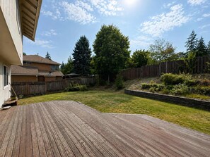 A backyard with a wooden deck, a grassy lawn, a wooden fence, and a house in the background. - Quiet Renton 3BR House near Valley Medical Ctr (Renton)