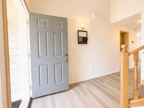 A hallway with a grey door, a wall-mounted light fixture, and a wooden staircase. - Quiet Renton 3BR House near Valley Medical Ctr (Renton)
