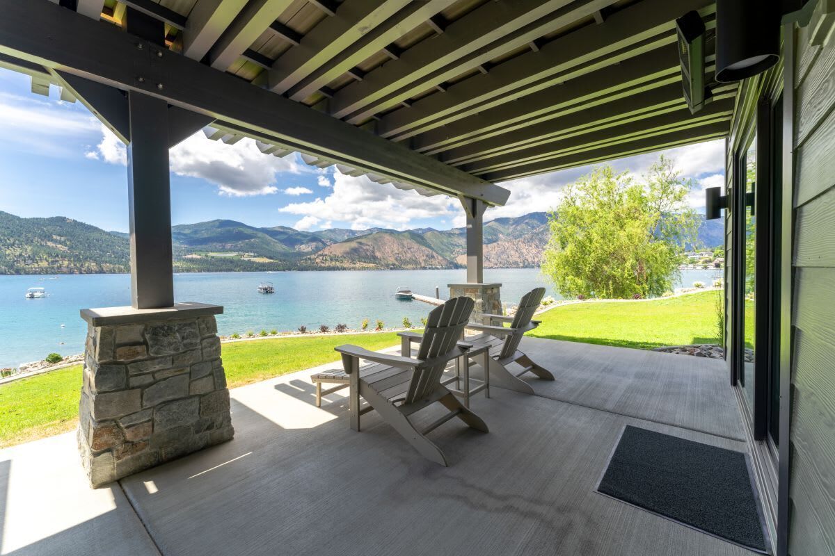 A covered patio with Adirondack chairs and a stone fireplace, overlooking a lake with boats and mountains.