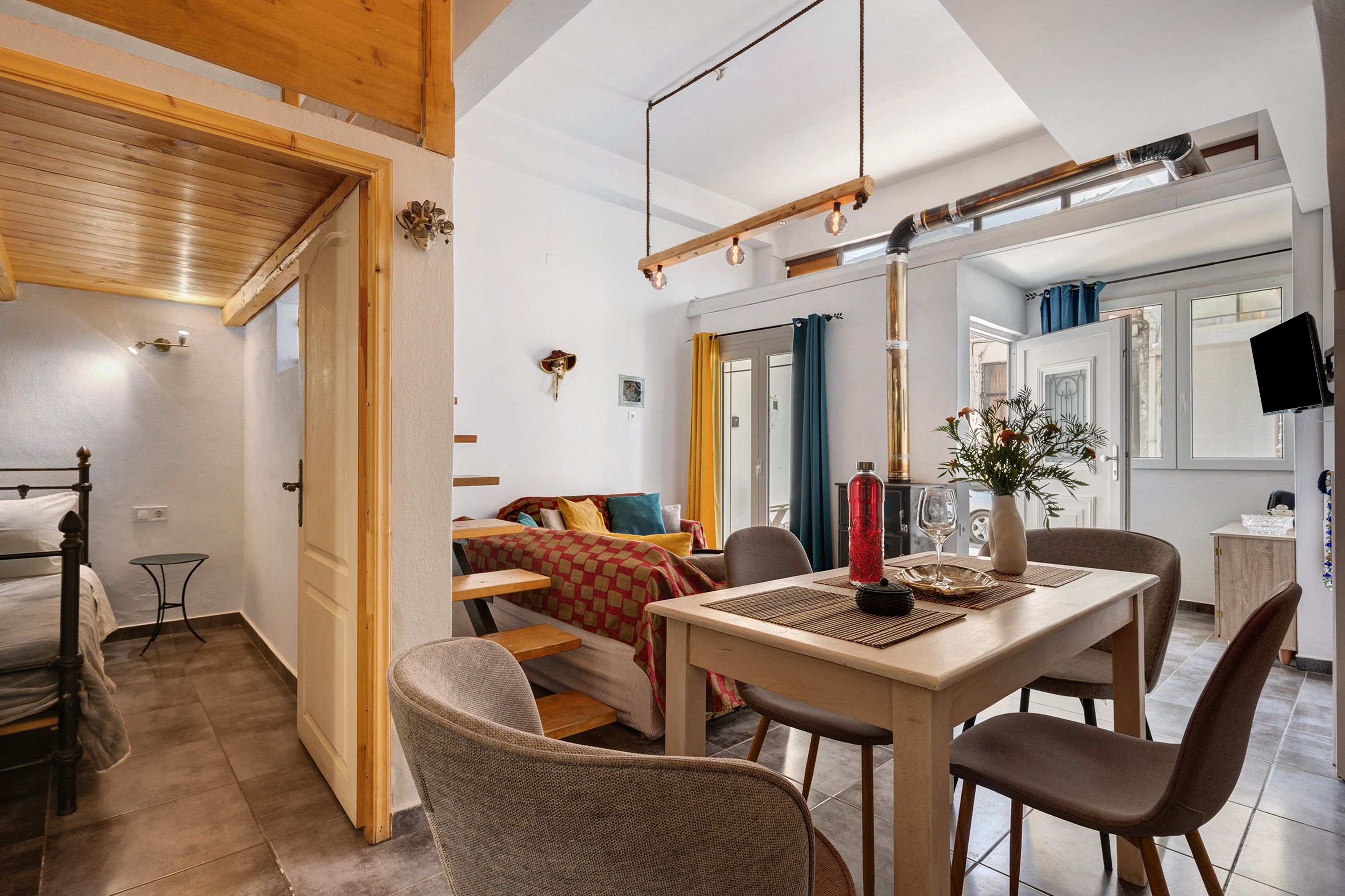 Un interior acogedor con una mesa de comedor de madera, sillas, una cama con ropa de cama colorida y una mesita con un florero y una botella.