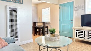 Un salon avec une table basse en verre, une porte bleue, un téléviseur posé sur un meuble et un panneau indiquant le mot « PLAGE ».