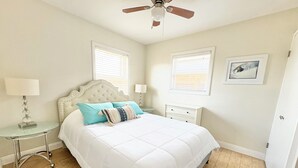 Une chambre à coucher comprenant un lit, une table de chevet, une lampe, un ventilateur de plafond et deux fenêtres équipées de stores.