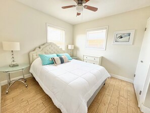 Ein Schlafzimmer mit einem Bett, einem Nachttisch, einer Lampe, einem Deckenventilator und zwei Fenstern mit Jalousien.