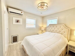 Een slaapkamer met een groot bed, een kroonluchter, twee ramen met zonwering en een luchtverkoeler aan de muur.