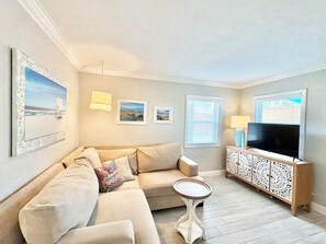 Een woonkamer met een bank, een klein rond tafeltje, een televisie op een kast en ingelijste foto's aan de muur.