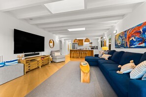 Ein modernes Wohnzimmer mit großem Flachbildfernseher, einer Couch, einem Couchtisch und einem Essbereich im Hintergrund.