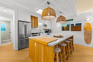 A modern kitchen with a central island, stainless steel appliances, wicker pendant lighting, and a surfboard decoration. - Perfect 4 Families, Walk To Beach, Roof-Top Patio! (Oxnard)