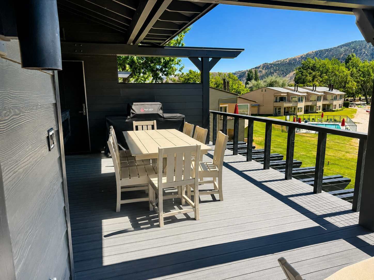 A covered outdoor deck with a table and chairs, a barbecue grill, and a view of a residential area with houses and a pool.