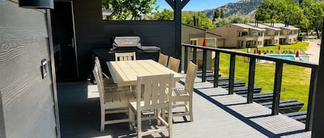 A covered outdoor deck with a table and chairs, a barbecue grill, and a view of a residential area with houses and a pool.