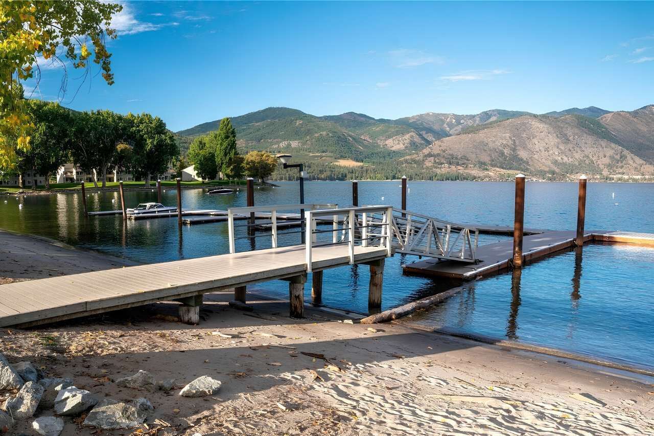 A lakeside dock with a wooden walkway leading to a docked boat, surrounded by mountains and trees.