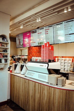 Mostrador de cafetería con una pizarra de menú encima, una máquina de café y pilas de vasos desechables.