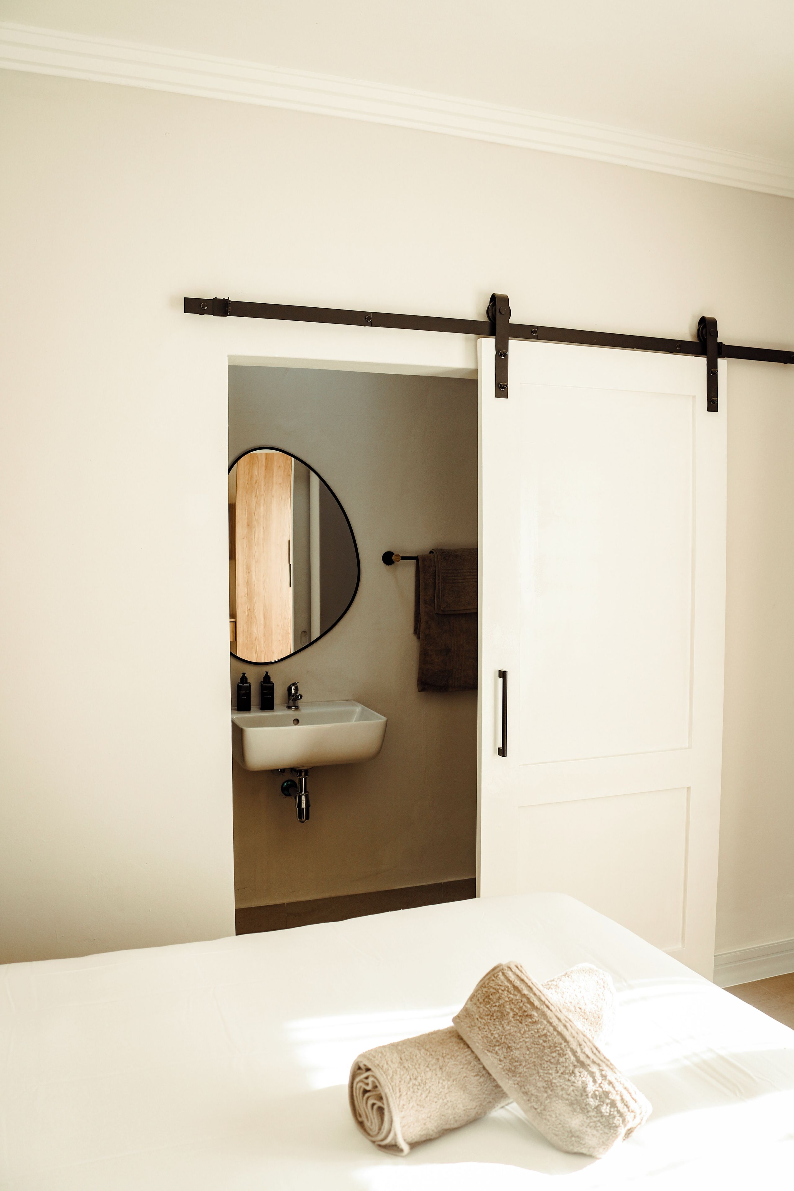 Habitación de hotel con cama, pared blanca, baño con lavamanos y espejo, y puerta corredera.
