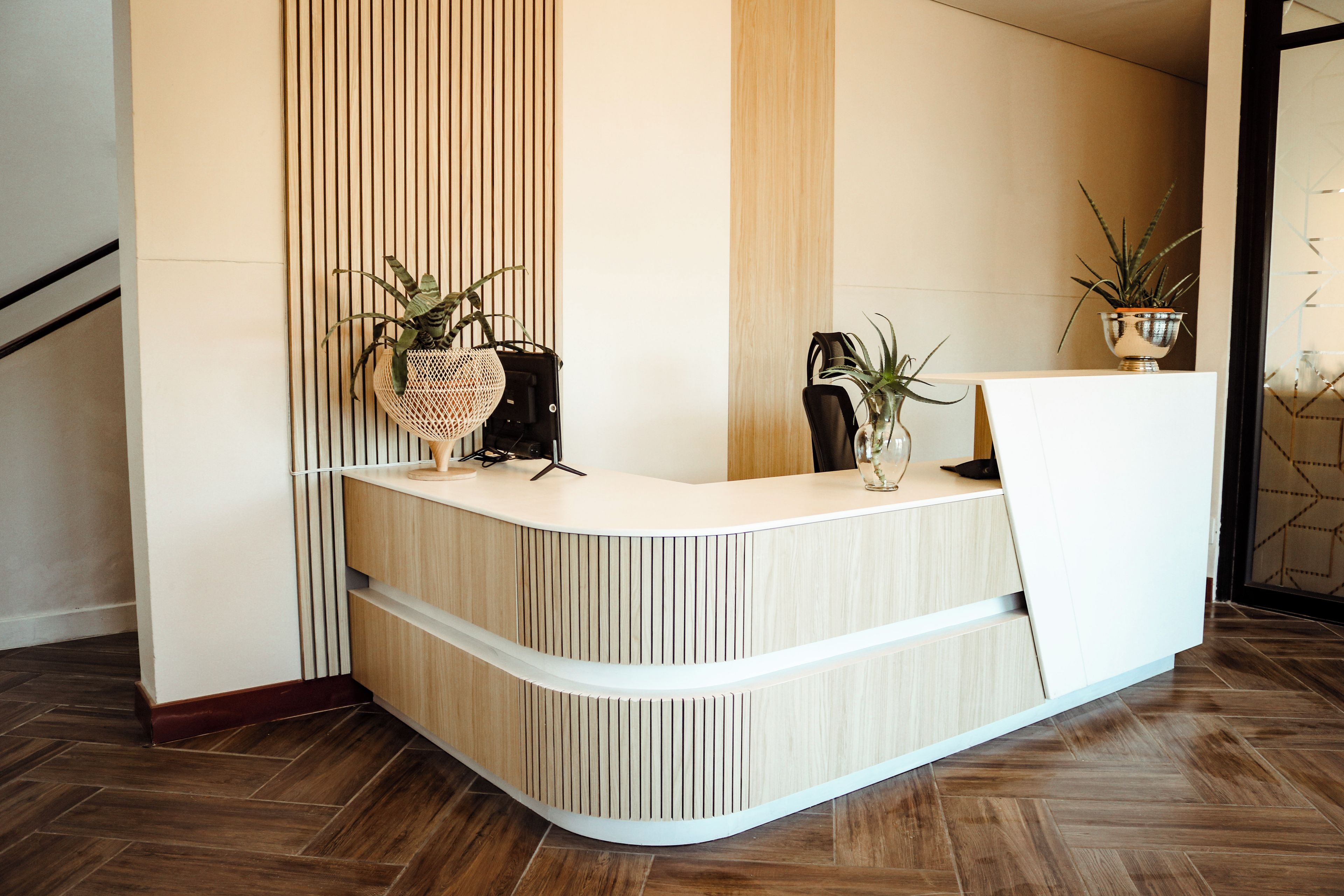 Un mostrador de recepción con listones verticales de madera, dos plantas en macetas y una silla moderna.