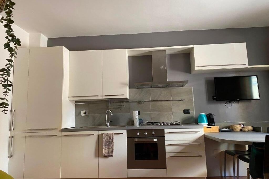 Una cucina moderna con mobili bianchi, una cappa aspirante in acciaio inossidabile, un forno a incasso e un televisore a schermo piatto montato a parete.